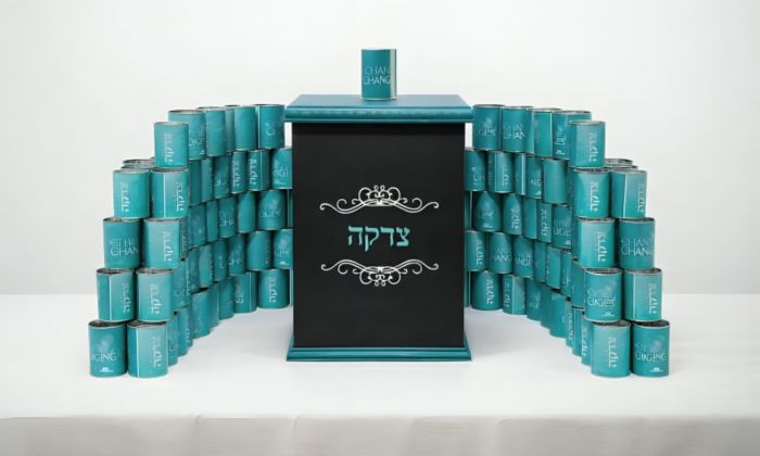 Teal-colored tzedakah (charity) box made of stacked cans arranged in a pyramid shape, with the word 'Tzedakah' written in Hebrew on the front. The cans have the text 'CHANGE CHANGES' printed on them, and the overall structure resembles modern art in stall