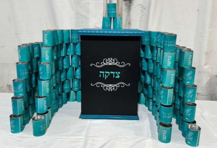 Teal-colored tzedakah (charity) box made of stacked cans arranged in a pyramid shape, with the word 'Tzedakah' written in Hebrew on the front. The cans have the text 'CHANGE CHANGES' printed on them, and the overall structure resembles a modern art instal